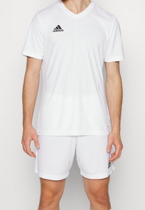 Mężczyzna ubrany w białą odzież sportową Adidas z krótkim rękawem i szortami, stoi z ramionami rozluźnionymi wzdłuż ciała na jednolitym tle.