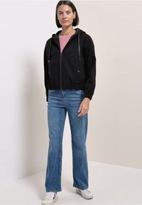 Felpa nera con zip e cappuccio, con vita elasticizzata e coulisse, abbinata a jeans a zampa chiara e sneakers bianche. Sfondo semplice.