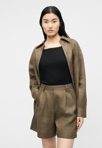 Γυναίκα που φοράει υπερμεγέθη πουκάμισο σε ελαιώδες πράσινο με αντίστοιχα σορτς ψηλής μέσης και μαύρο αμάνικο τοπ, στέκεται με τα χέρια στις τσέπες.