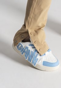 Białe sneakersy z niebieskimi akcentami i dużym napisem "GIANNI". Połączone z beżowymi spodniami z detalami zatrzaskowymi na końcu nogawek.