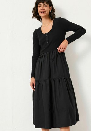 Schwarzes Kleid mit enganliegendem Langarm-Oberteil mit Rundhalsausschnitt und Knöpfen, das in einen gestuften, plissierten Rock übergeht. Glatte Textur.