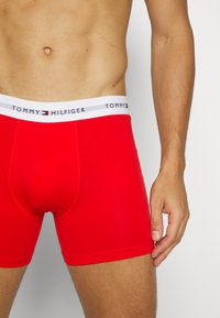 Rde bombažne boksarice z belim pasom, na katerem je v črnih črkah napis "TOMMY HILFIGER". Mehka, oprijeta zasnova.