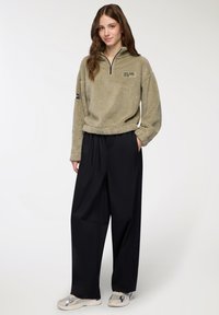 Młoda kobieta w beżowym polarze z napisem "New York Athletics Club", czarnych szerokich spodniach i beżowych sneakersach, stojąca z jedną ręką w kieszeni.