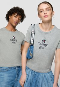 Pilkos, trumpomis rankovėmis marškinėliai su "tommy girl" tekstu ir žvaigždės logotipu; moteris vilki mėlyną klostuotą džinsinę sijoną ir maišelį su žaviomis detalėmis.