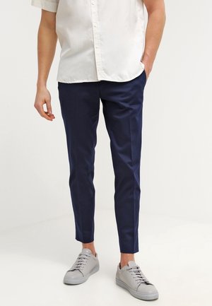 Mężczyzna ubrany w granatowe, dopasowane spodnie, białą koszulę na krótki rękaw z guzikami oraz jasnoszare sportowe buty, stoi z jedną ręką w kieszeni.