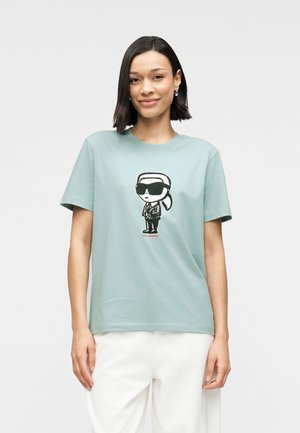 Ženska s kratkimi temnimi lasmi, oblečena v svetlo modro majico z risanim moškim v sončnih očalih in belimi hlačami, stoji ob belem ozadju.