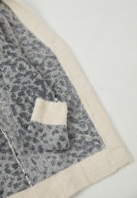 Mjukt stickat tyg med ett grått och svart leopardmönster samt krämfärgade kanter och manschetter liggande platt på en vit yta.