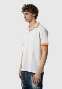 Polo bianco con colletto arancione e accenti sulle maniche. Presenta una piccola placca e un logo ricamato sul petto. Indossati jeans in denim sotto.