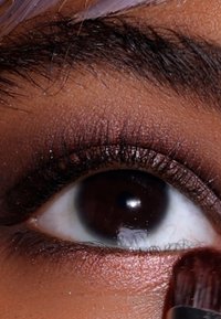 Primer plano de un ojo marrón con una sombra de ojos bronce brillante aplicada, pestañas finas visibles y un pincel de maquillaje en contacto con la piel.