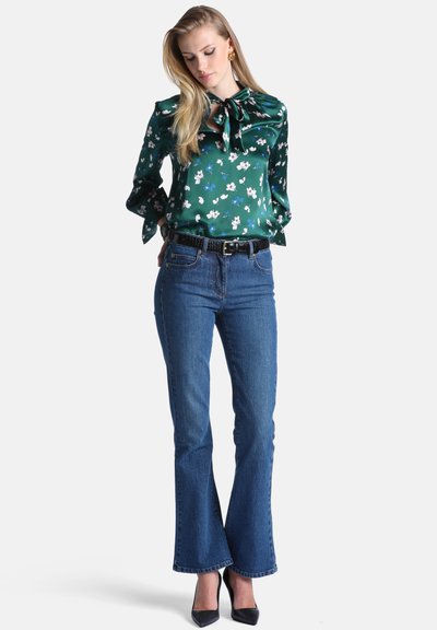 Femme portant un chemisier vert à fleurs à manches longues avec un nœud, un jean évasé bleu taille haute, une ceinture noire et des talons noirs pointus, debout les mains dans le dos.