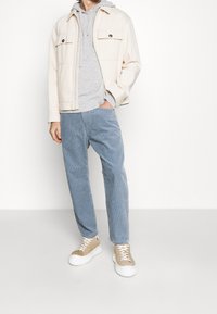 Giacca in corda beige con due taschini sul petto indossata sopra un hoodie grigio. Pantaloni in corda dritti azzurro chiaro e sneakers beige con suole bianche.