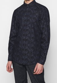 Camicia blu navy con bottoni, caratterizzata da un motivo testuale tonale, maniche lunghe e colletto strutturato, realizzata in un tessuto liscio.