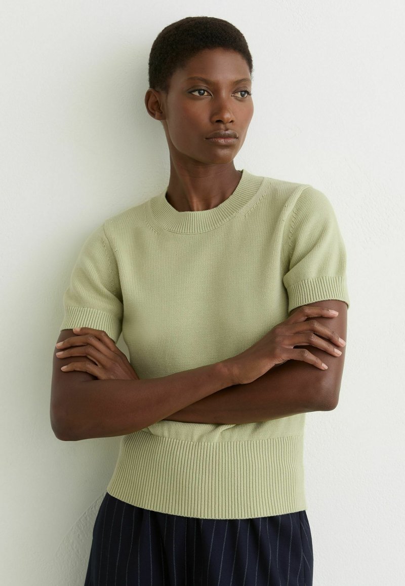 Femme aux cheveux courts portant un haut en tricot vert clair à manches courtes et un pantalon rayé sombre, bras croisés, debout contre un mur blanc.