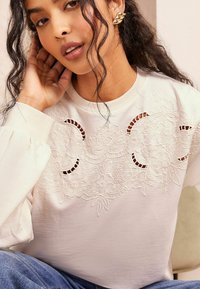 Lys beige sweatshirt med indviklet blomsterbroderi og udskæringsdetaljer. Lange, puffede ærmer og en rund halsudskæring fuldender designet.