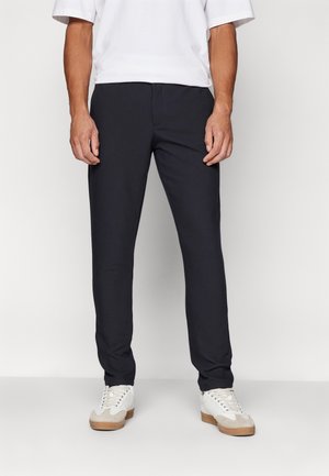 Homme portant un pantalon noir slim-fit, des baskets blanches et une chemise blanche à manches courtes, debout devant un fond clair uni.