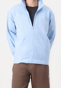 Ljusblå zip-up jacka med texturerad yta, utrustad med två framfickor och en liten logotyp på bröstet, tillsammans med bruna byxor.