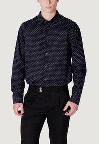 Camicia nera a maniche lunghe con pois bianchi, design con bottoni, tessuto morbido, vestibilità su misura. Indossata con pantaloni neri plissettati.