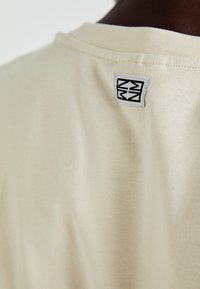 Beige pamut póló, amelyen a hátsó nyakkivágás és egy geometrikus fekete mintájú varrott címke látható. Sima textúrájú, klasszikus fazon.