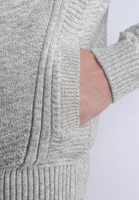 Harmaa neulepusero, jossa on teksturoitu neulekuvio, ribbaatut hihat ja sivutasku. Kangas näyttää pehmeältä ja paksulta, mikä luo kotoisan ilmeen.