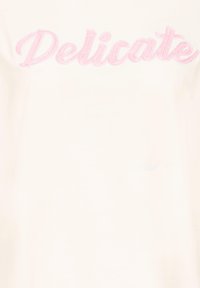 Camiseta blanca con texto bordado en rosa que dice "Delicate". Tela suave, mangas cortas y cuello redondo. Diseño sencillo.