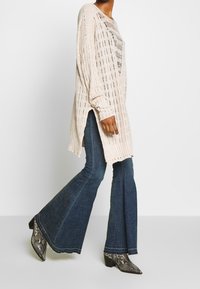 Un pull en tricot beige clair avec un tissage ouvert, doté de manches longues et de fentes sur les côtés, associé à un jean foncé évasé et des bottines imprimé serpent.