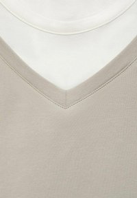 Tricouri v-neck stratificate; unul în bej deschis cu țesătură texturată și unul dedesubt din bumbac alb, prezentând culori și cusături contrastante.