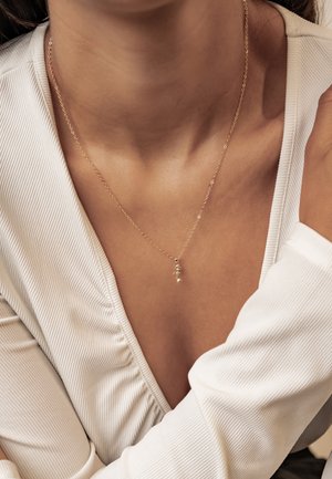 Gold chain necklace with a small pendant worn around the neck over a white ribbed V-neck top.
