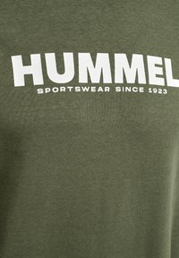Camiseta de algodón verde oliva con un bold logo en blanco de "HUMMEL" y el texto "ROPA DEPORTIVA DESDE 1923". Textura suave, corte casual.
