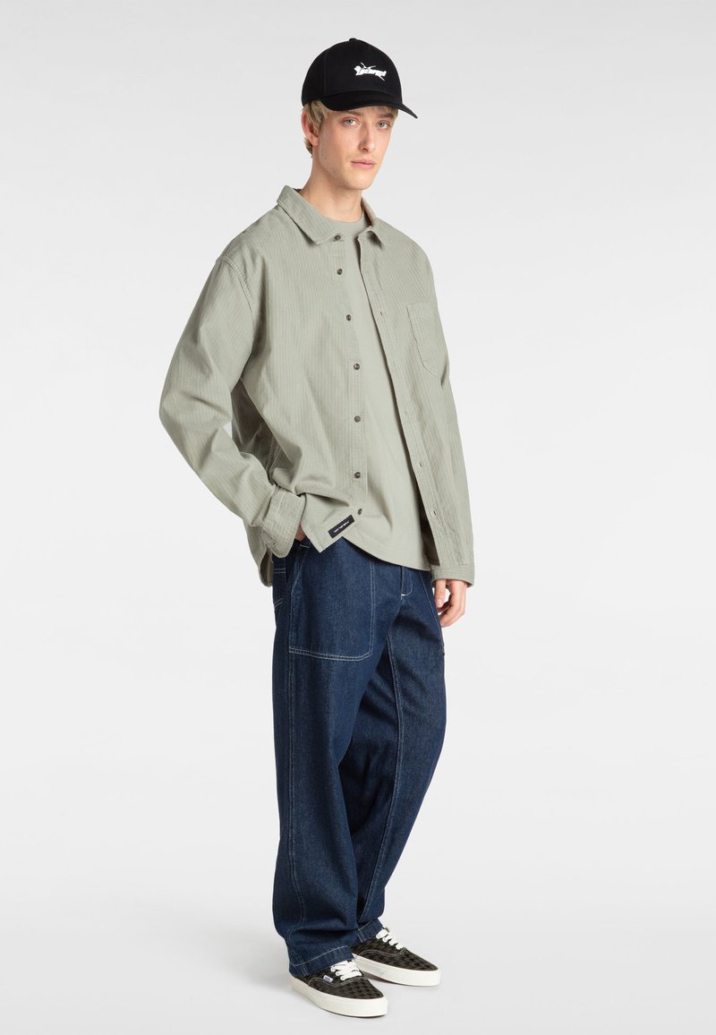 Lichtgroene geribde button-up shirt over een grijze T-shirt, gecombineerd met loszittende blauwe denim broek en zwarte geblokte sneakers. Zwarte pet op het hoofd.