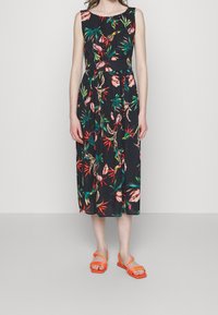 Blumenkleid in Midi-Länge mit tailliertem Oberteil aus dunklem Stoff, versehen mit pinken und grünen tropischen Mustern, kombiniert mit orangefarbenen Riemchensandalen.