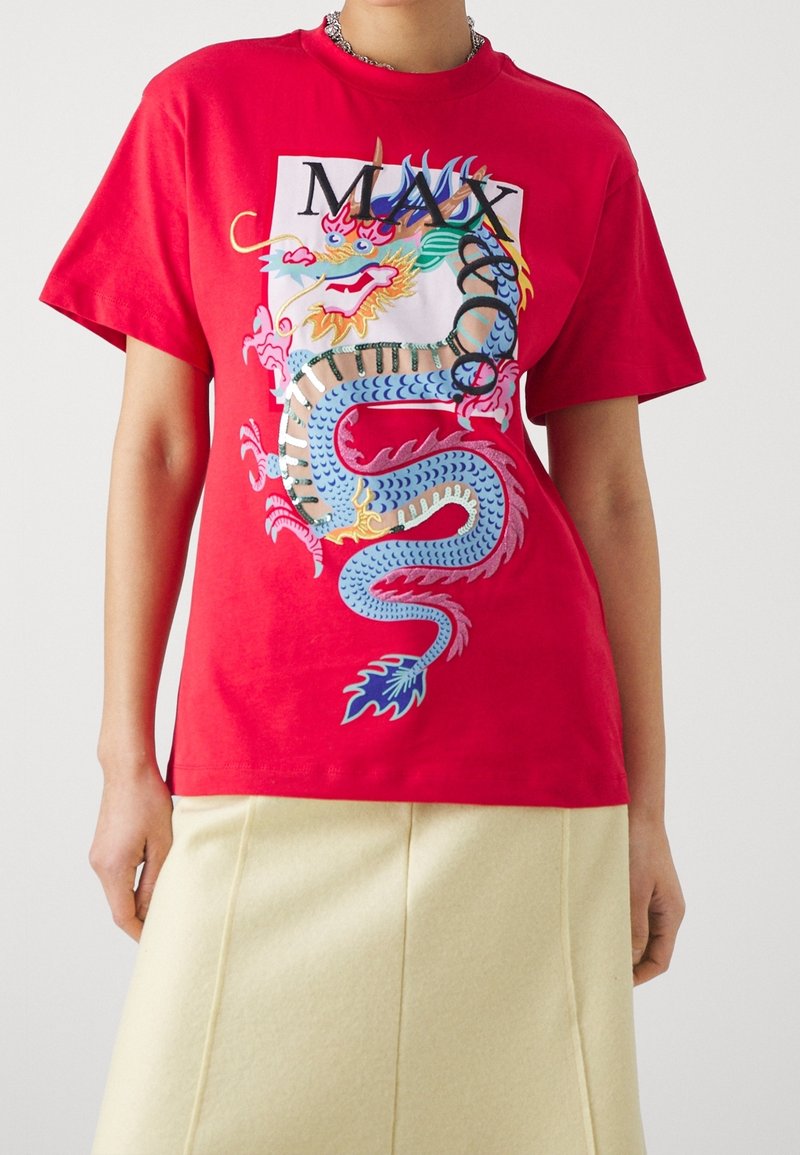 Personne portant un t-shirt rouge avec un dragon brodé coloré et le texte "MAX", associé à une jupe beige clair sur un fond uni.