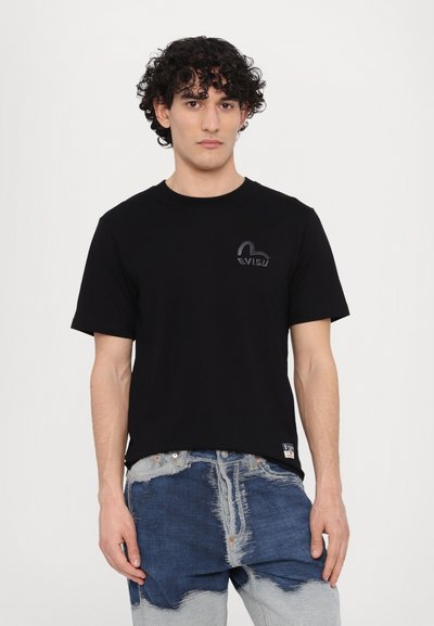 T-shirt noir à manches courtes avec un col rond, arborant un logo subtil en violet foncé sur la poitrine. Le modèle l'associe à un jean en denim déchiré.