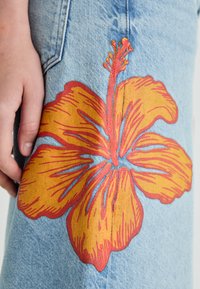 Peça de vestuário em denim com um grande bordado de flor hibisco em laranja e vermelho. Textura subtil no tecido com cor de denim azul. Mão humana visível.
