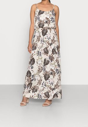 Kvinde iført en ærmeløs creme maxi-kjole med brune og grønne tropiske blad- og blomsterprint, kombineret med beige sandaler med åben tå.