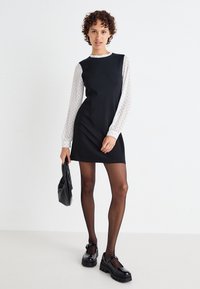 Musta hihaton mekko, jonka alla on teksturoitu valkoisilla pilkuilla varustettu pitkähihaiseen paita. Malli pitää mustaa laukkua, ja hänellä on päällään opakimmat mustat sukkahousut ja jykevät kengät.