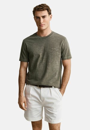 Man draagt een olijfgroene korte mouwen t-shirt en witte geplooide shorts met een zwarte riem, staand met één hand in de zak.