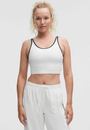 Weißes Crop-Top mit marineblauer Einfassung, mit rundem Ausschnitt und dünnen Trägern, kombiniert mit lockeren weißen Jogginghosen.