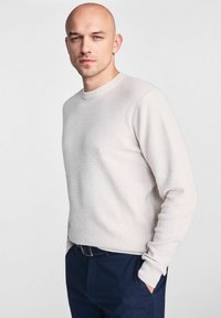 Jasno-beżowy sweter z prążkowanego materiału, z długimi rękawami i okrągłym dekoltem, w zestawieniu z ciemnoniebieskimi spodniami. Tkanina wydaje się teksturowana i dopasowana.