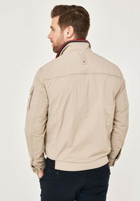Beige jacket with a structured collar, featuring contrasting red and navy stripes. Side pocket and ribbed waistband. Smooth fabric texture.