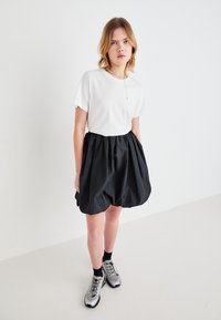 Witte T-shirt met korte mouwen gecombineerd met een zwarte, knielange, volumineuze rok. De outfit wordt aangevuld met zilveren sneakers en zwarte sokken.