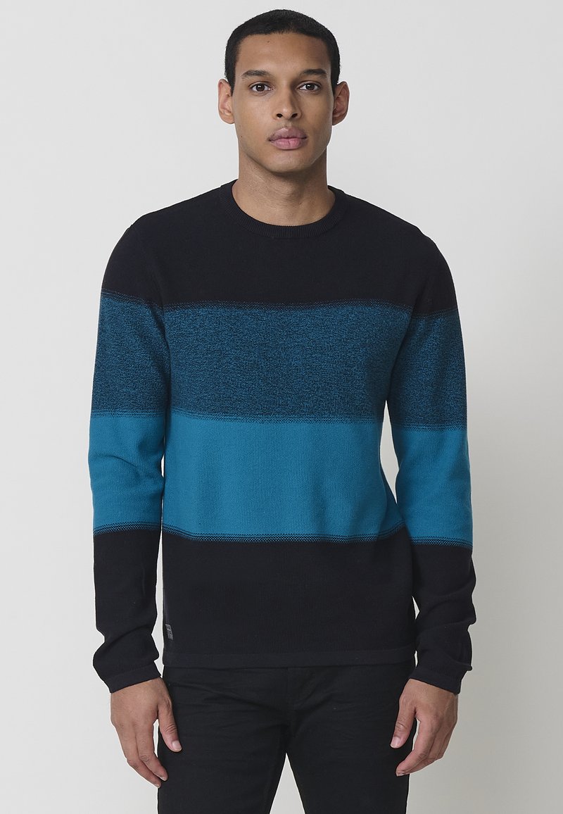 Adīta džemperis ar melnu augšējo daļu, teksturētu tējā zilo vidējo joslu un vienkrāsainu tējā zilo apakšējo daļu. Apaļa kakla izgriezuma forma un garas piedurknes.