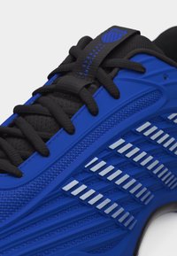 Μπλε αθλητικό παπούτσι με μαύρα κορδόνια, επάνω μέρος από δίχτυ, λευκές ανακλαστικές ρίγες και μαύρη ετικέτα με το λογότυπο στην γλώσσα.