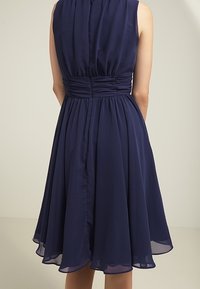 Sukienka bez rękawów w granatowym kolorze w kształcie litery A z marszczoną talią i swobodną spódnicą do kolan, pokazana od tyłu na neutralnym tle.