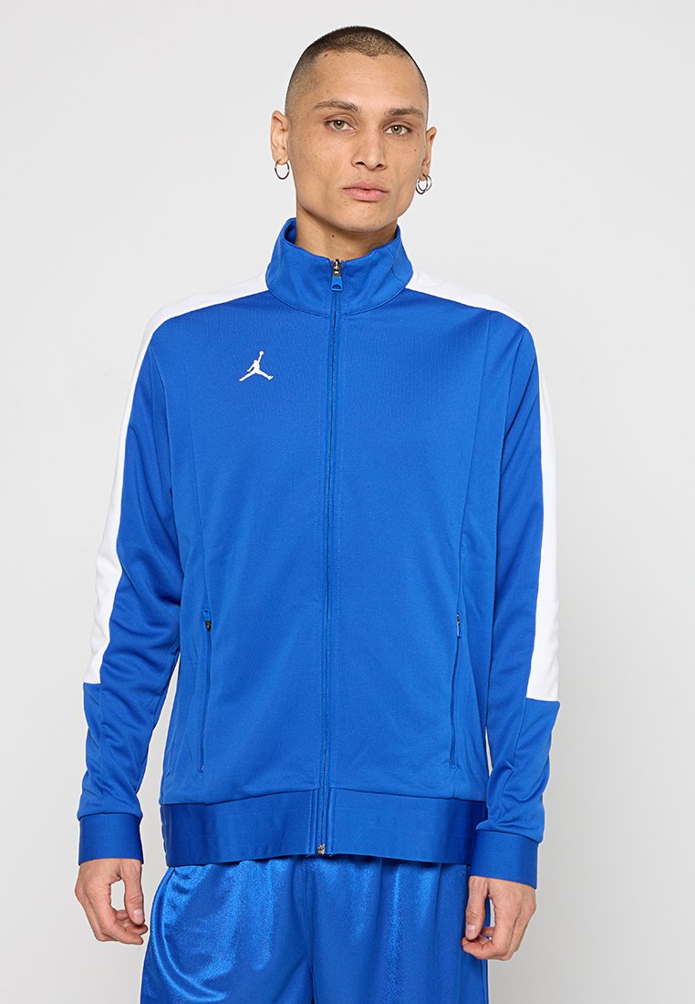 Blue athletic jacket with white side stripes, high collar, zip closure, and embroidered logo on the chest. Smooth, breathable fabric.