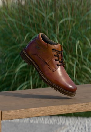 Shoe de couro castanho com bico redondo, atacadores e colar texturizado. Apresenta uma sola robusta e um acabamento suave, posicionado acima de uma superfície de madeira.