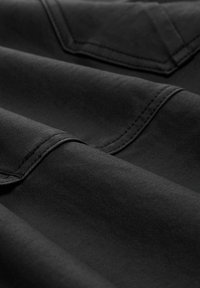 Tissu noir avec coutures détaillées et vue partielle de deux poches angulaires, montrant la texture et les lignes de couture.