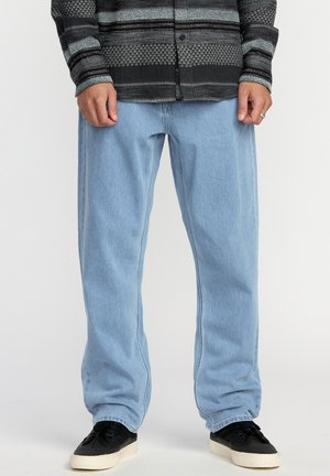 Homme portant un jean ample bleu clair, des baskets noires et blanches, et une chemise manches longues à motifs noirs et gris, debout devant un fond blanc.