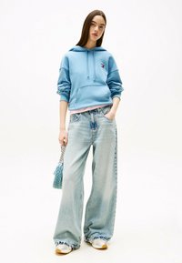 Sudadera con capucha de color azul claro, de corte holgado, con bolsillo tipo canguro. Pantalones vaqueros de pierna ancha en azul claro con dobladillos deshilachados. Zapatillas blancas y un bolso pequeño.