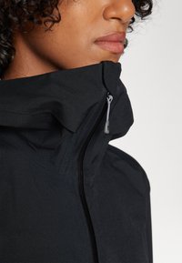 Black jacket with a high collar and a zip closure. Features a textured fabric and a drawstring detail on the collar.