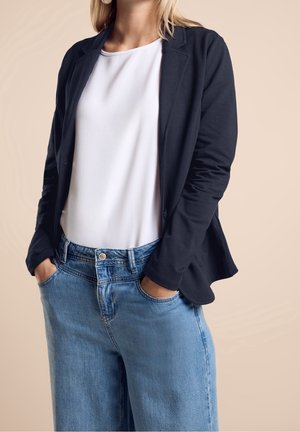 Vrouw draagt een zwarte blazer over een wit topje, met blauwe spijkerbroek, handen in de zakken, staand tegen een effen beige achtergrond.
